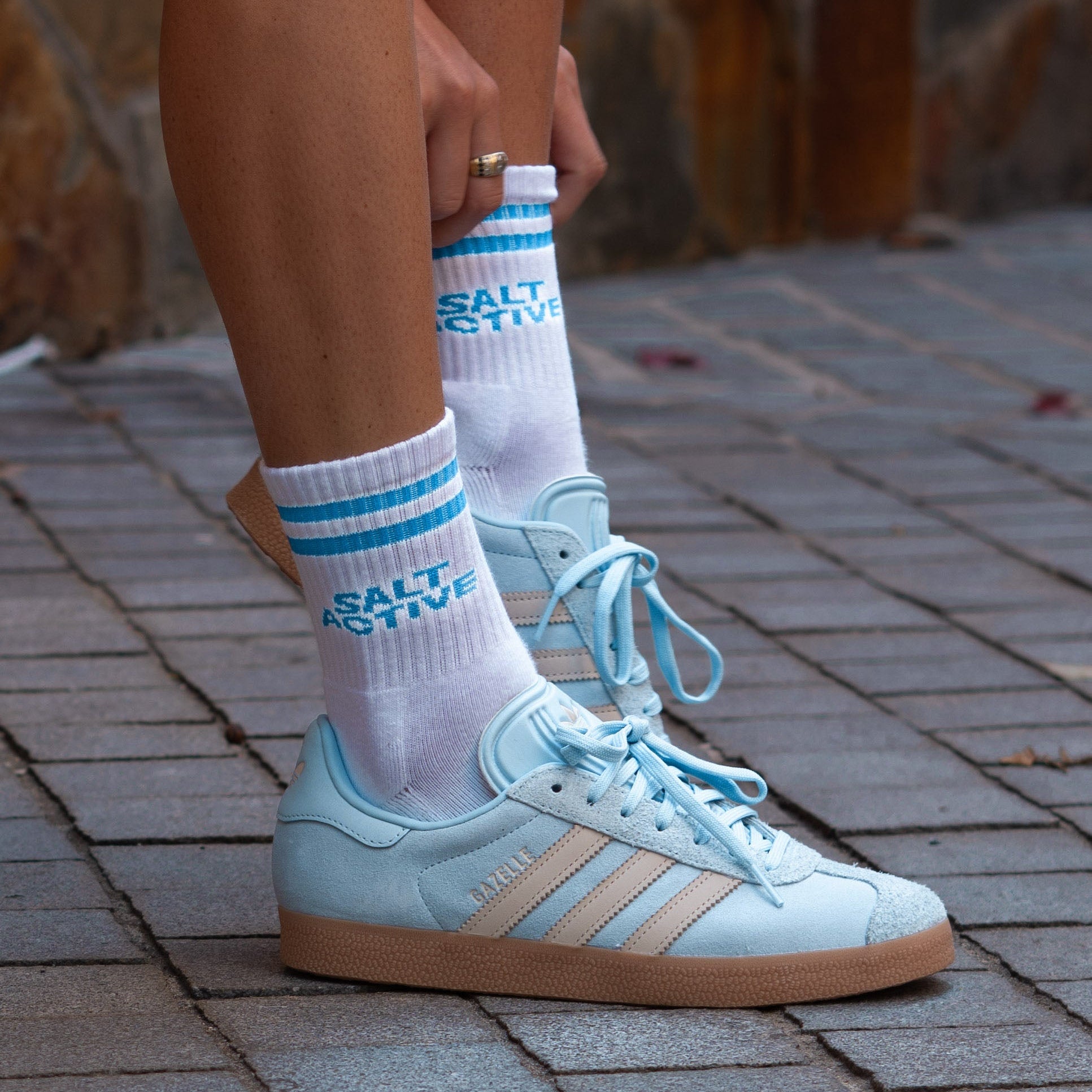 Person wearing light blue Adidas sneakers with white socks featuring text, standing on a brick pavement.